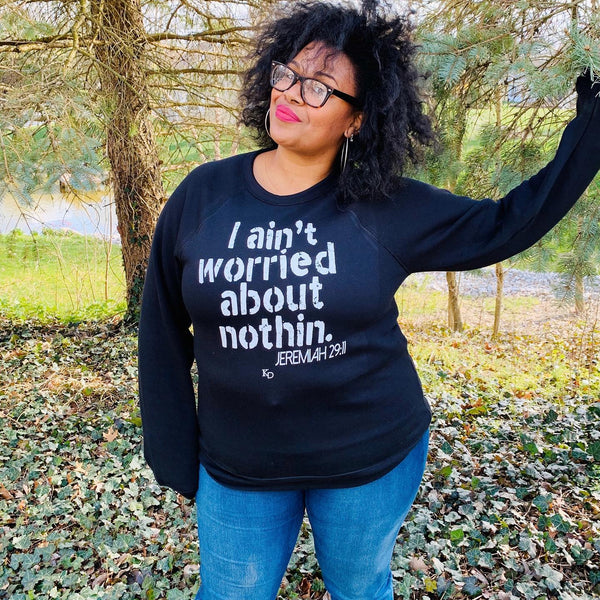 black woman in glasses and red lipstick wearing black soft pullover sweatshirt bella and canvas with screen print design saying i aint worried about nothin referencing jeremiah 2911 bible verse